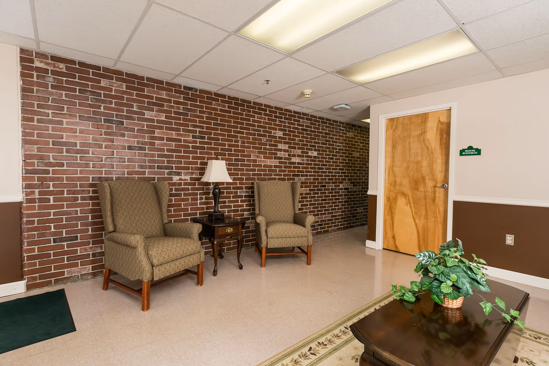 Small waiting area with two upholstered armchairs flanking a side table and lamp against a brick accent wall, a wooden door, and a coffee table with a potted plant.