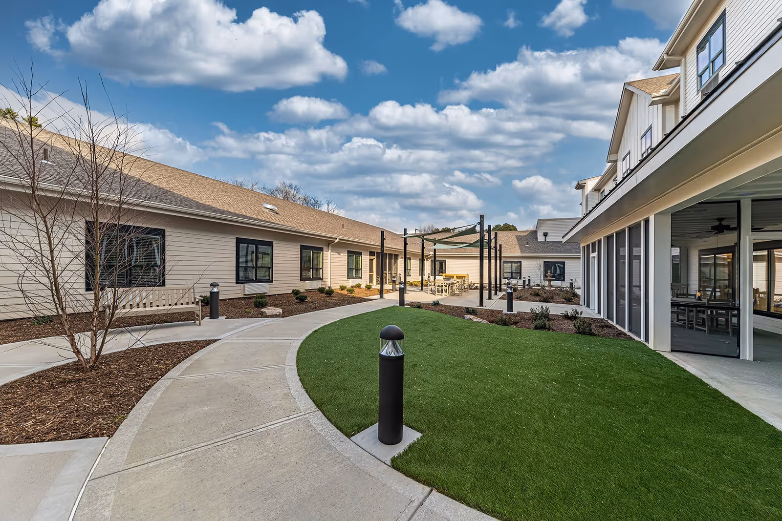 Outdoor courtyard area at Cloverland Park Senior Living featuring a curved concrete walkway, green lawn, benches, and a shaded seating area with tables and chairs. The surrounding buildings have beige siding and multiple windows under a partly cloudy blue sky.