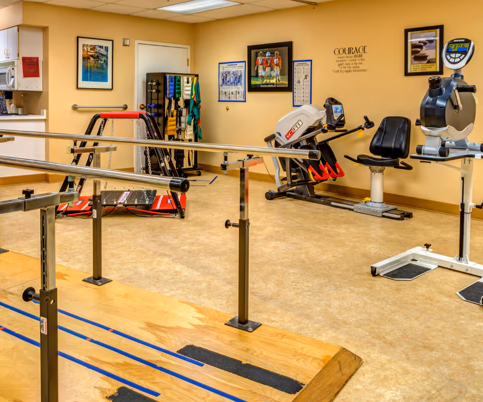 A rehabilitation or physical therapy room with parallel bars for walking exercises, exercise equipment including a seated leg press machine and a stationary arm bike, resistance bands hanging on a rack, and motivational posters on the walls.