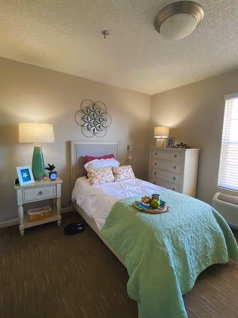 A cozy bedroom with a single bed covered in white and green bedding, decorated with multiple pillows. There is a nightstand with a lamp, a framed photo, and a small plant on the left side of the bed. On the right side, there is a tall dresser with a lamp and decorative items on top. A window with blinds allows natural light into the room. The walls are painted beige, and a decorative metal wall art piece hangs above the bed.