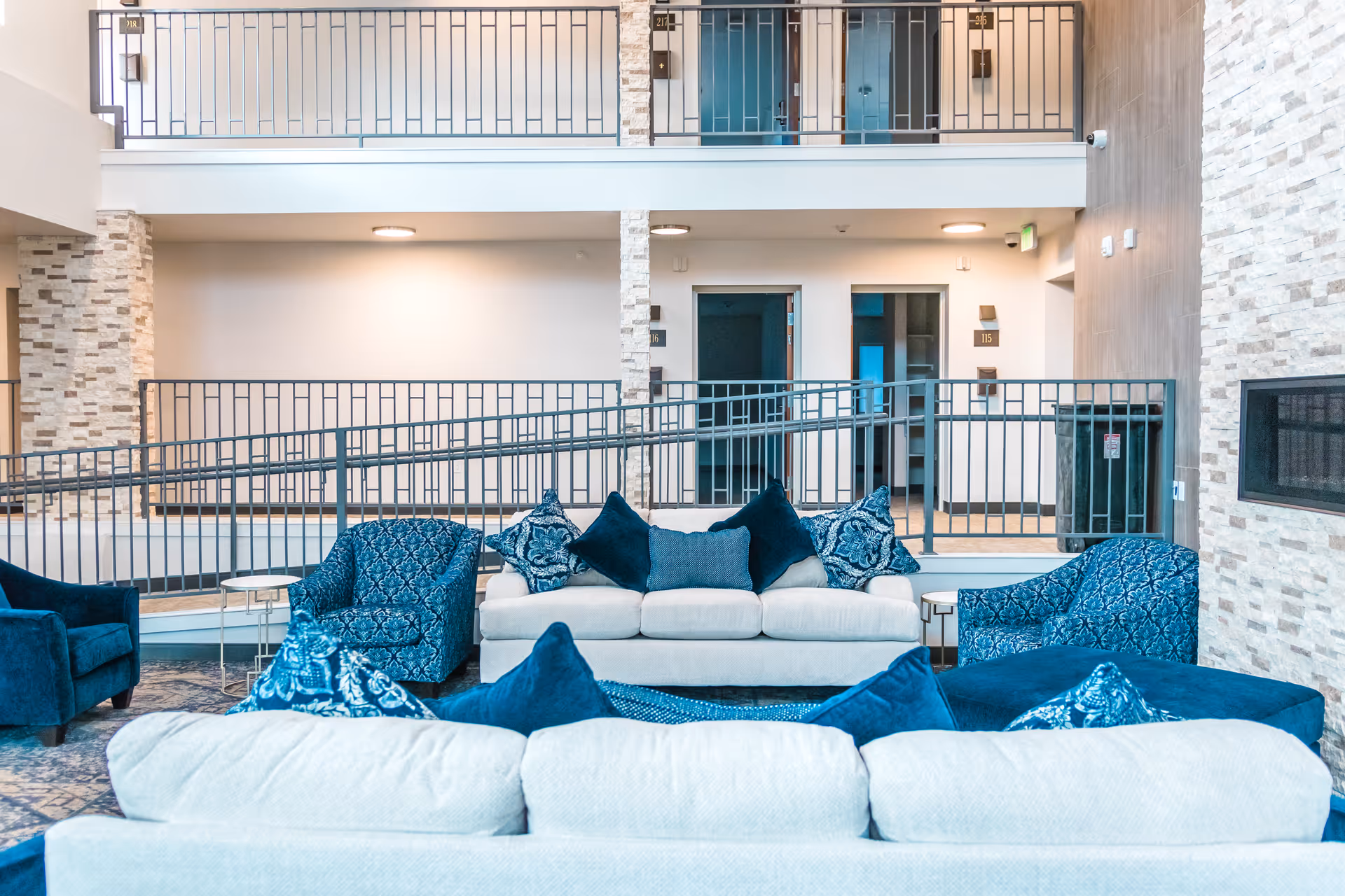 A modern indoor common area in Downtown Assisted Living featuring two beige sofas with blue patterned and solid blue pillows, two blue patterned armchairs, and a blue ottoman. The space has a stone accent wall with a built-in fireplace on the right, metal railings, and doors to rooms in the background.