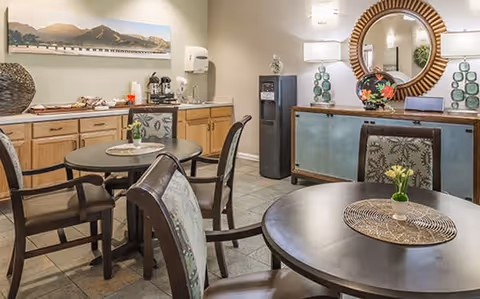 A cozy dining area with round tables and cushioned chairs. Each table has a decorative placemat and a small flower vase. In the background, there is a wooden sideboard with a round mirror above it, two lamps, and decorative items. On the left side, there is a counter with coffee and snack supplies, and a water dispenser is visible near the corner.
