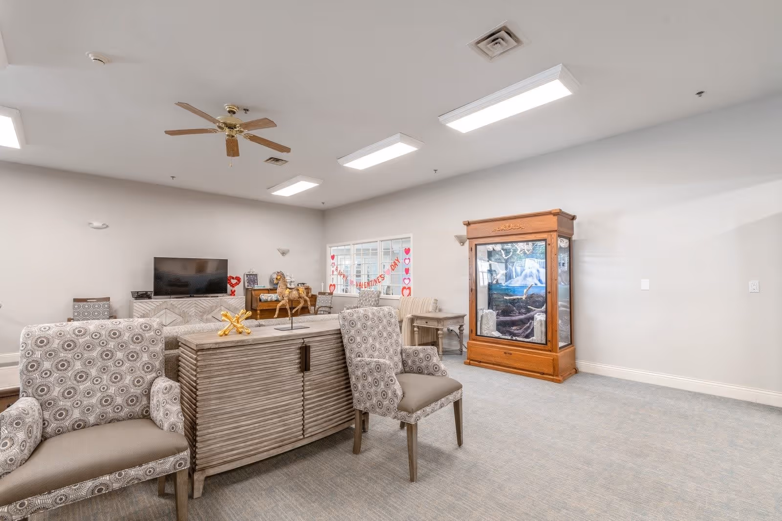 A bright and spacious living room area in a senior living facility with patterned armchairs, a wooden cabinet, a TV on a stand, a ceiling fan, and a large aquarium. The room has light-colored walls and carpeted flooring, with Valentine's Day decorations visible on the window.
