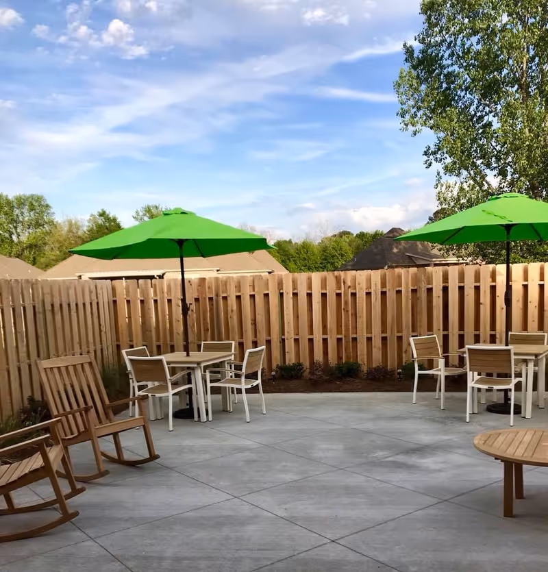 Outdoor patio area with wooden rocking chairs, tables with white chairs, and green umbrellas. The space is enclosed by a wooden fence with trees and rooftops visible in the background under a partly cloudy sky.