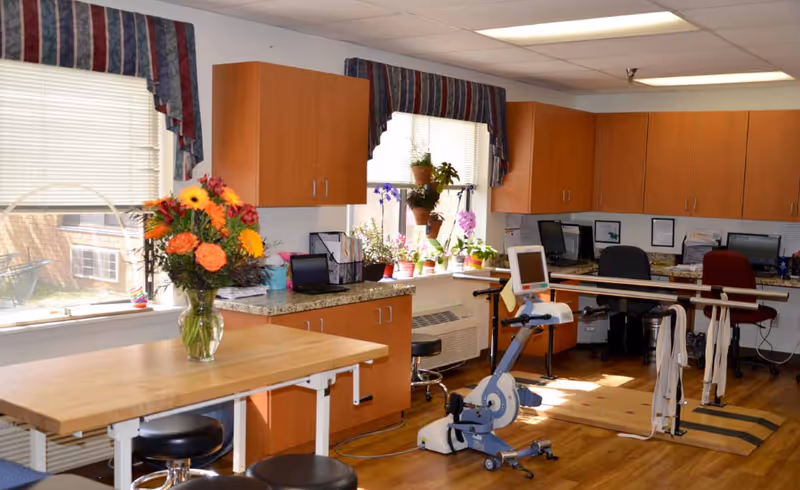 A bright room with wooden cabinets and countertops, a table with a vase of colorful flowers, several potted plants on the windowsill, two office chairs at desks with computers, and physical therapy equipment including a stationary bike and parallel bars.