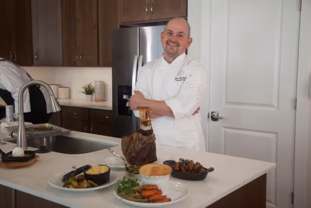 A chef in a white uniform stands smiling with arms crossed behind a kitchen island displaying a large cooked tomahawk steak, a bowl of mashed potatoes, roasted vegetables, and a plate of cooked carrots and green beans in a modern kitchen with wooden cabinets and a stainless steel refrigerator.