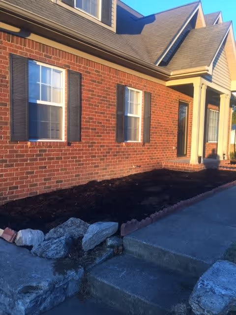 Exterior view of a brick building with two windows featuring black shutters and a front door with a small porch. There is a garden bed with dark soil bordered by rocks and bricks, and a concrete walkway with steps leading up to the porch.