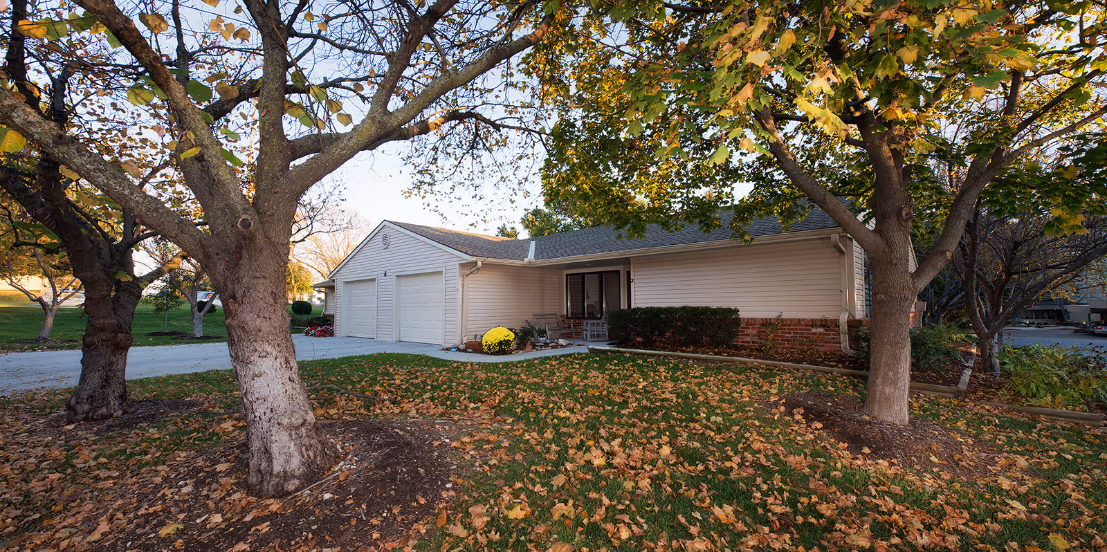 Exterior view of a single-story residential building with a three-car garage, surrounded by trees with autumn leaves on the ground and green grass.