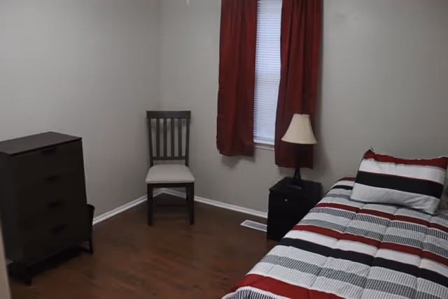 A small bedroom with a single bed covered in a striped bedspread in red, black, white, and gray. There is a wooden chair with a white cushion against the wall, a dark wooden dresser with three drawers, and a small black nightstand with a lamp. A window with red curtains is on the wall behind the chair.