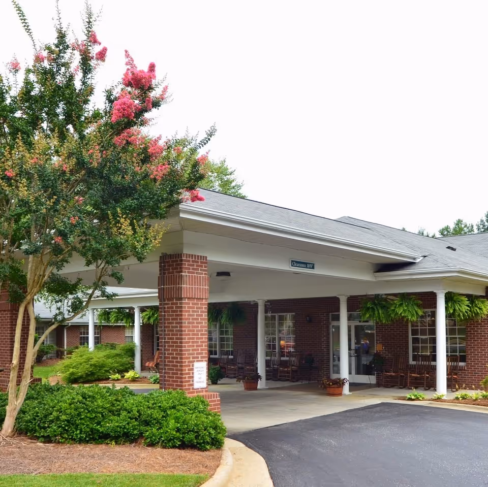 Entrance of a senior living facility with a covered drop-off area supported by white columns and brick pillars. There are rocking chairs and potted plants under the covered area, with green shrubs and a tree with pink flowers in the foreground.