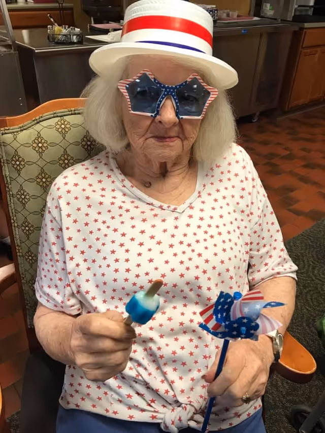 An elderly woman wearing a white shirt with red stars, star-shaped sunglasses with an American flag pattern, and a white hat with red and blue stripes. She is sitting in a chair indoors, holding a blue and white popsicle in one hand and a small American flag-themed pinwheel in the other.