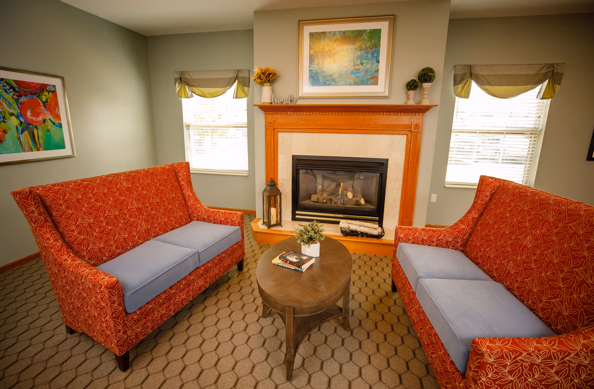 A cozy living room with two red patterned sofas with blue cushions facing each other across a round wooden coffee table. The table has a small potted plant and two books on it. Behind the table is a fireplace with a wooden mantle decorated with a framed colorful painting, a vase with flowers, and small topiary plants. Two windows with beige and green valances flank the fireplace, and a colorful abstract painting hangs on the left wall.