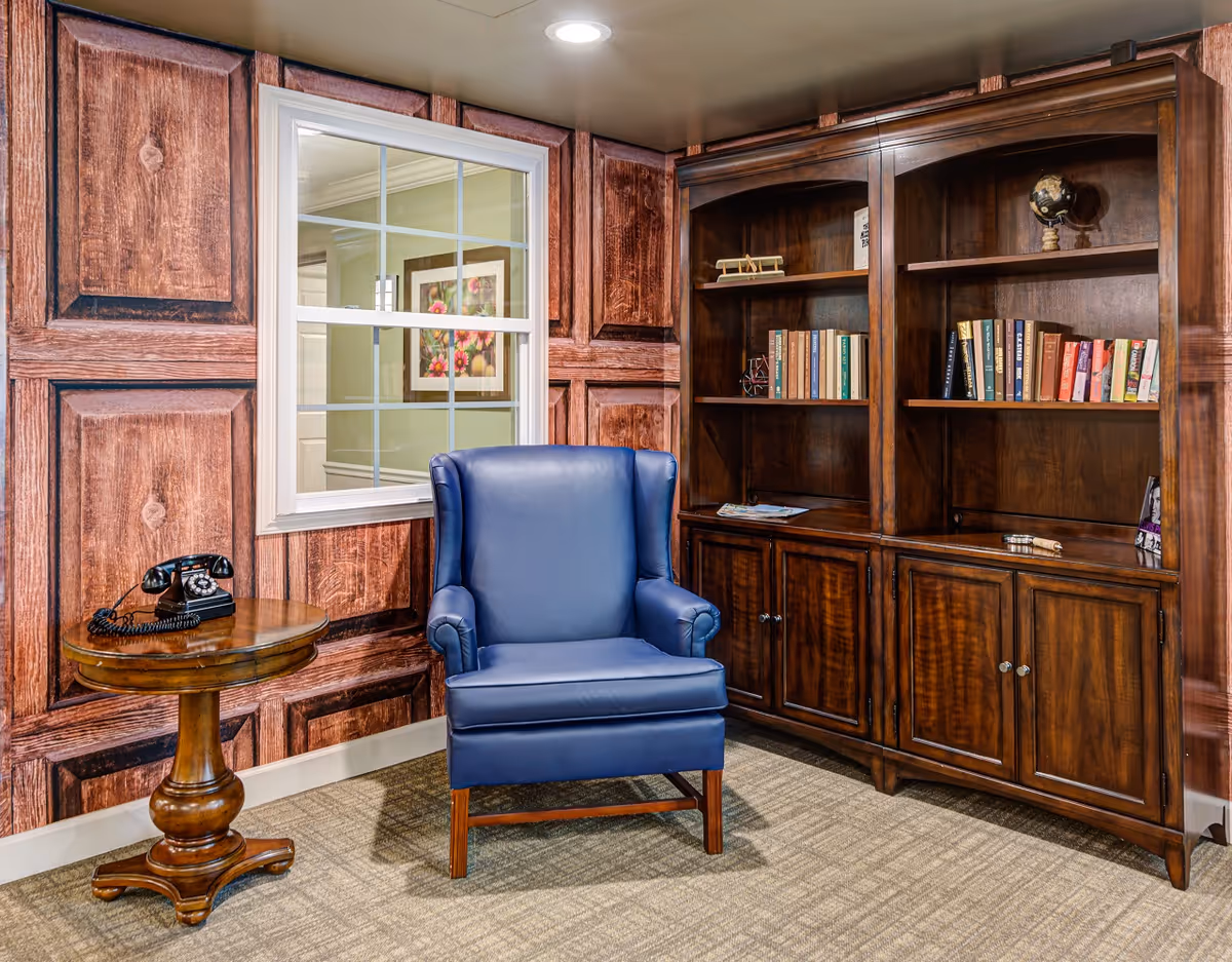 A blue leather armchair sits beside a round wooden side table with a vintage telephone, in front of wood-paneled walls and a large wooden bookcase filled with books.