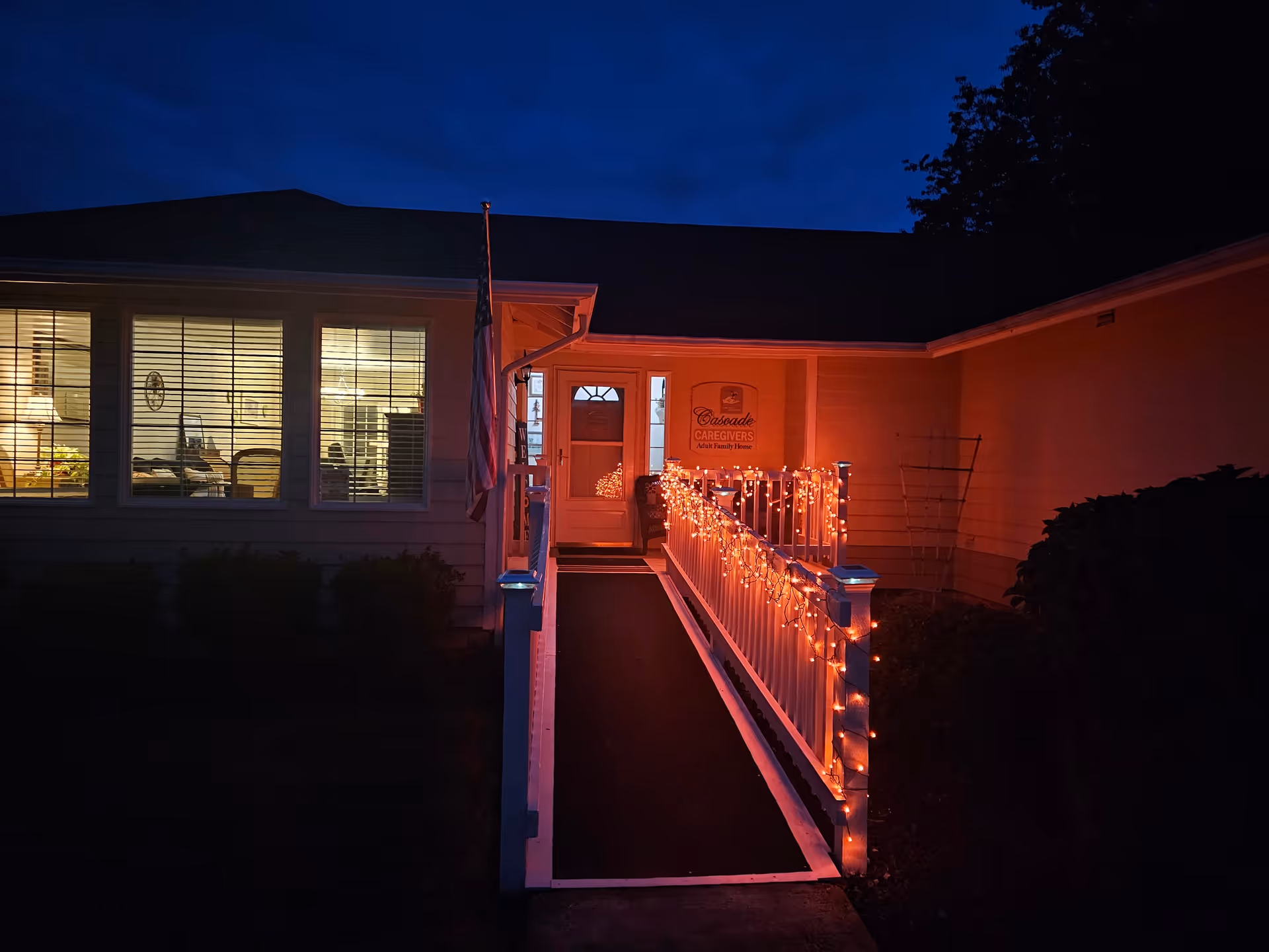 Night view of the entrance to Cascade Caregivers Adult Family Home with a ramp decorated with orange string lights. The building is lit from inside, and an American flag is displayed near the entrance.
