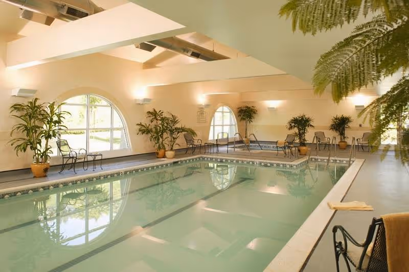 Indoor community swimming pool with arched windows, potted plants, seating, and exposed ceiling beams.