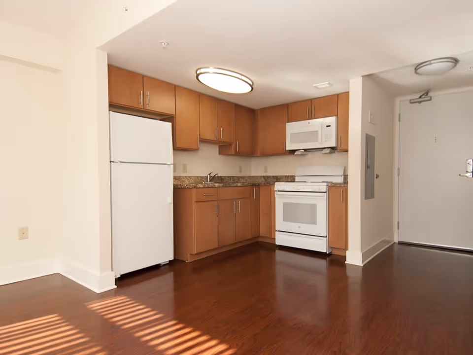 A small kitchen area with wooden cabinets, a white refrigerator, a white stove with oven, and a white microwave above the stove. The kitchen has a granite countertop and dark wood flooring. There is a white door to the right of the kitchen area and a ceiling light fixture above.