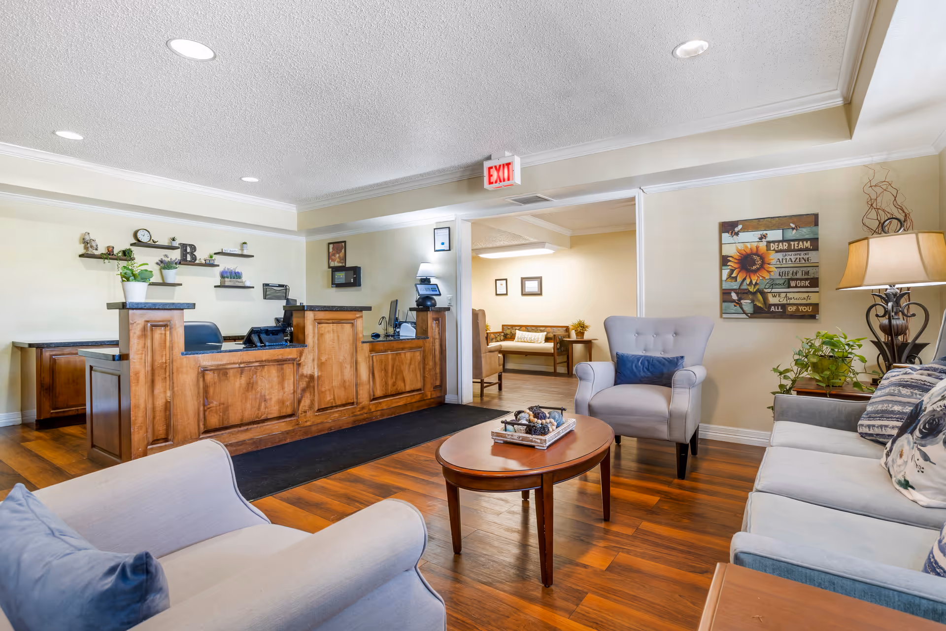 Reception area of Brookdale Memorial City featuring a wooden front desk with office equipment, a seating area with a light gray armchair, a blue-gray sofa with patterned pillows, a wooden coffee table with decorative items, and a lamp on a side table. The walls are light-colored with framed artwork and plants, and there is an exit sign above the doorway leading to another room with additional seating.
