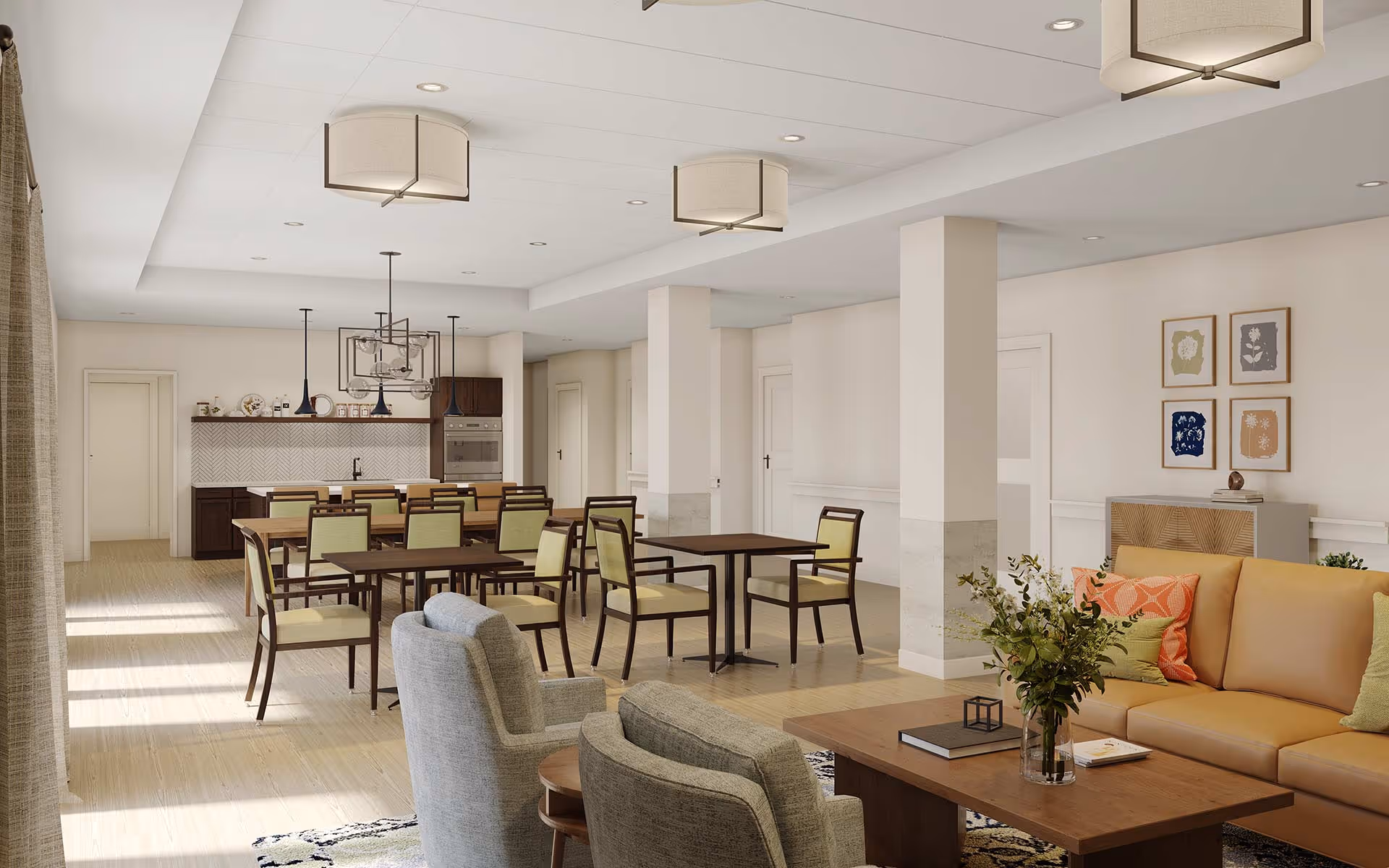 A bright and spacious common area in a senior living facility featuring a seating area with a tan sofa and gray armchairs around a wooden coffee table with a vase of flowers. In the background, there is a dining area with multiple tables and chairs, and a kitchen area with dark cabinetry and modern appliances. The room has light-colored walls, wooden flooring, and ceiling lights.
