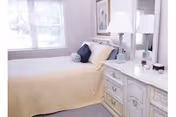 A cozy bedroom featuring a neatly made bed with a beige bedspread, a dark blue bolster pillow, and a framed picture on the wall above the bed. Next to the bed is a white dresser with a lamp, a mirror, and some decorative items. A window with white curtains allows natural light to brighten the room.