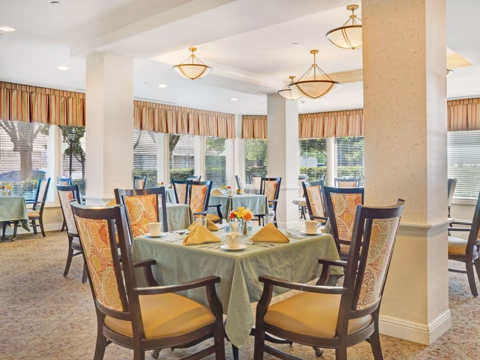 A bright dining room with multiple tables covered in green tablecloths, each set with yellow folded napkins, white cups, and small flower arrangements. The room has large windows with striped valances, patterned carpet, and elegant wooden chairs with patterned upholstery. Ceiling lights hang above the tables.
