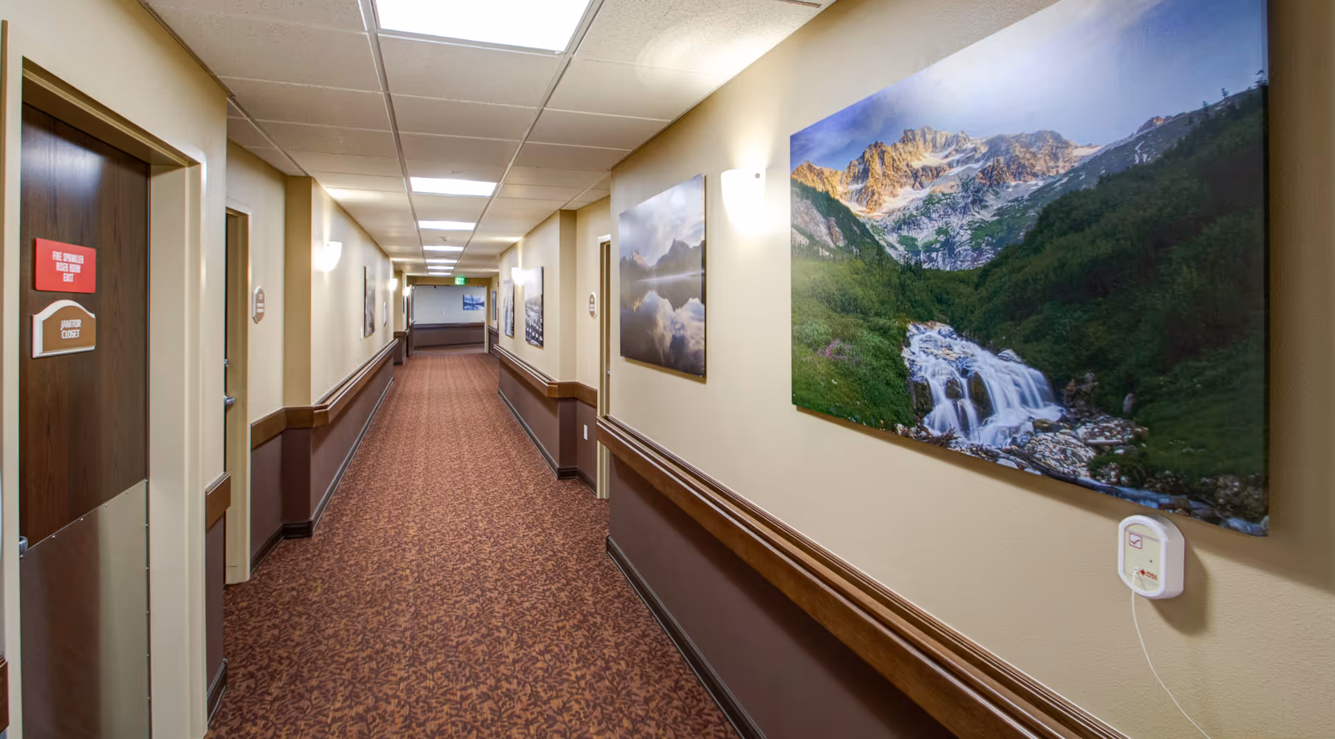 A long indoor hallway with patterned carpet and beige walls, decorated with large landscape photographs of mountains and waterfalls. The hallway has several doors and is well-lit with ceiling lights and wall sconces.