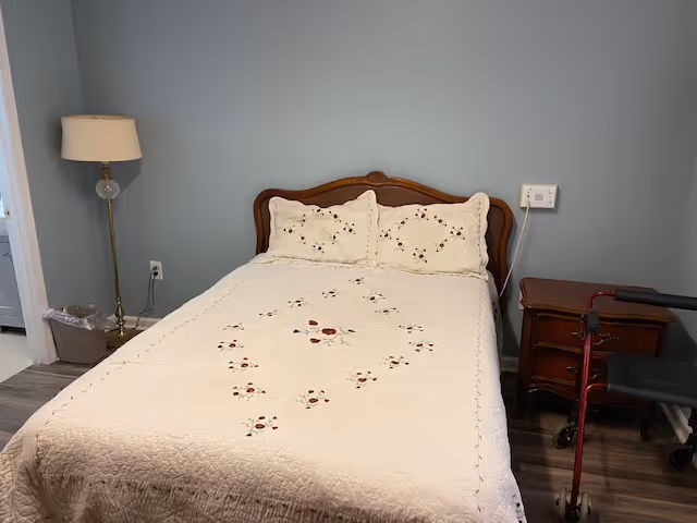 A small bedroom with a neatly made bed covered in a white quilted bedspread, a floor lamp and wastebasket on the left, a nightstand on the right, and a red walker beside the bed.