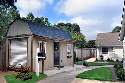 A small beige shed with a black door and a window air conditioning unit on the side, situated next to a paved walkway with green grass patches and a wooden fence in the background. There is a mailbox on a white post near the shed and a wooden wheelbarrow on the grass. Another building with a window and plants is visible on the right side.