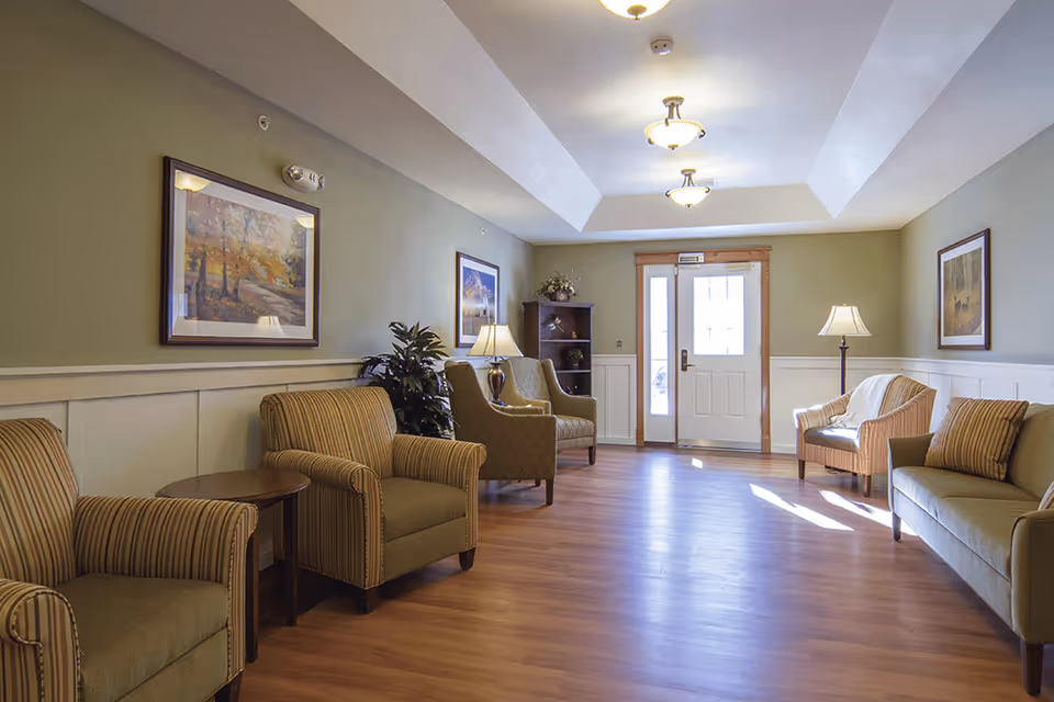 A well-lit living room area with wooden flooring, green walls, and white wainscoting. The room features several upholstered armchairs and a sofa arranged along the walls, two framed paintings, two table lamps, a floor lamp, a small wooden table, and a wooden shelving unit near a door with glass panels allowing natural light to enter.