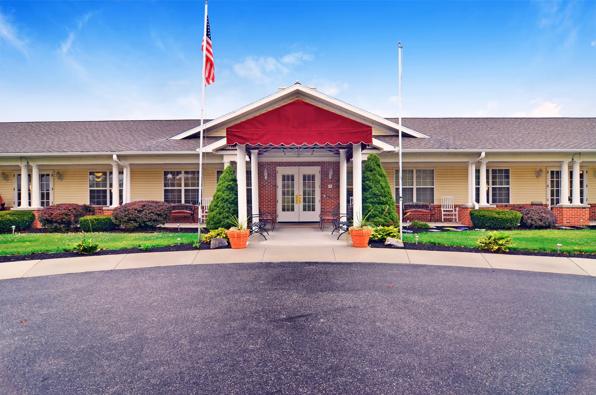 Front exterior view of Celebration Villa Of Shippensburg building with a covered entrance featuring a red awning, two flagpoles with an American flag, and landscaped greenery including bushes and potted plants.