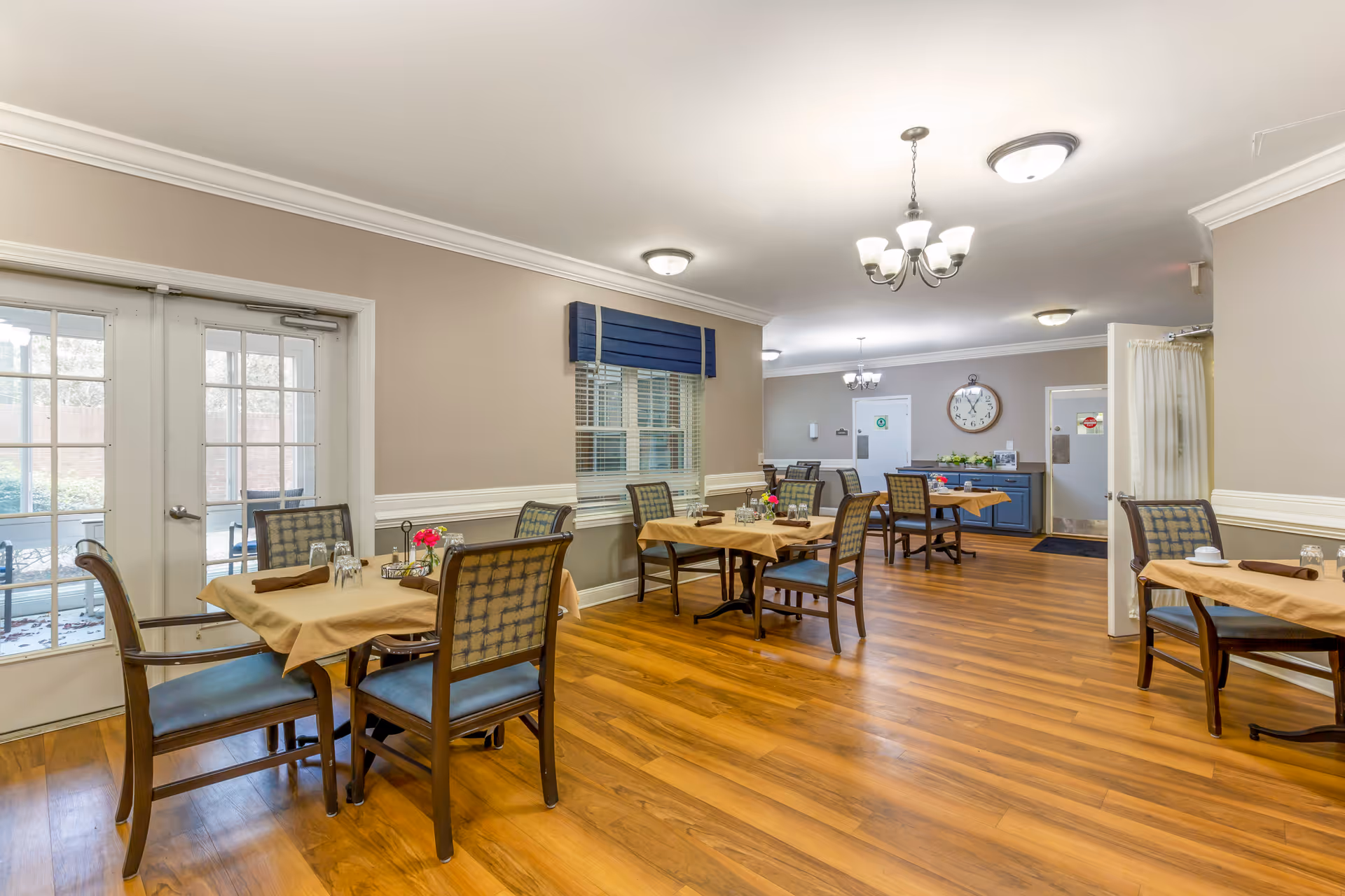 Well-lit communal dining room with multiple tables and chairs set with tablecloths and place settings.