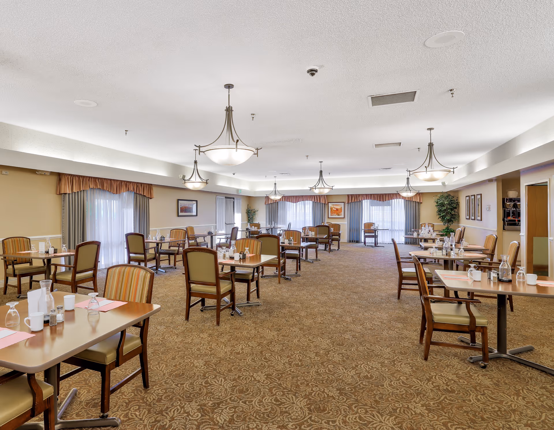 A spacious dining room with multiple tables and chairs arranged neatly. Each table is set with placemats, glasses, cups, and condiments. The room has carpeted flooring with a patterned design, beige walls, and large windows covered with curtains and valances. Several hanging light fixtures illuminate the room, and there are framed pictures and a potted plant along the walls.