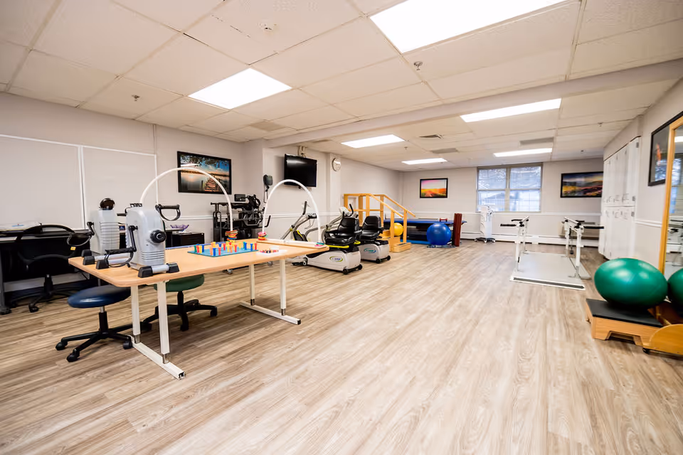 A spacious rehabilitation or physical therapy room with exercise equipment including stationary bikes, parallel bars, therapy balls, and a table with hand exercise devices. The room has wood-patterned flooring, white walls, fluorescent ceiling lights, and several framed pictures on the walls.