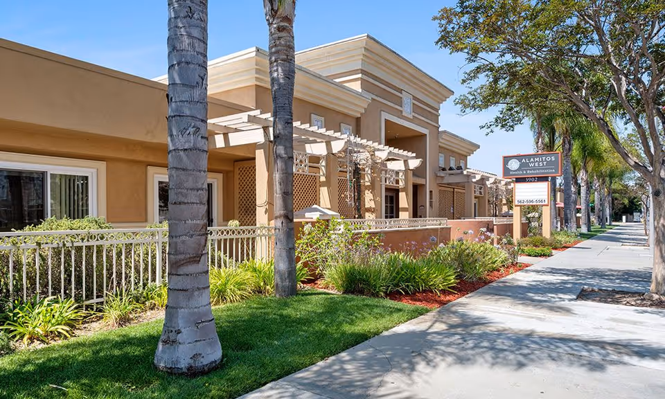 Front exterior of a single-story beige health and rehabilitation building with palm trees, landscaped beds, a sidewalk, and a sign for Alamitos West Health & Rehabilitation.