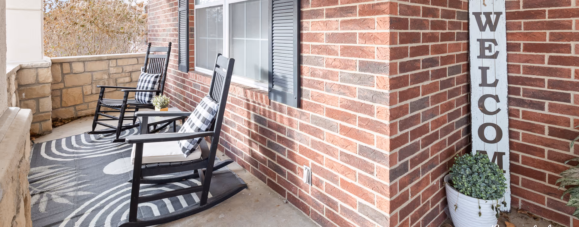 A cozy outdoor porch area with two black rocking chairs, each with a black and white checkered cushion. The porch has a patterned black and white rug on the floor, a small table with a potted plant between the chairs, and a tall vertical wooden sign leaning against a brick wall that reads 'WELCOME'. There is also a white planter with green foliage next to the sign.