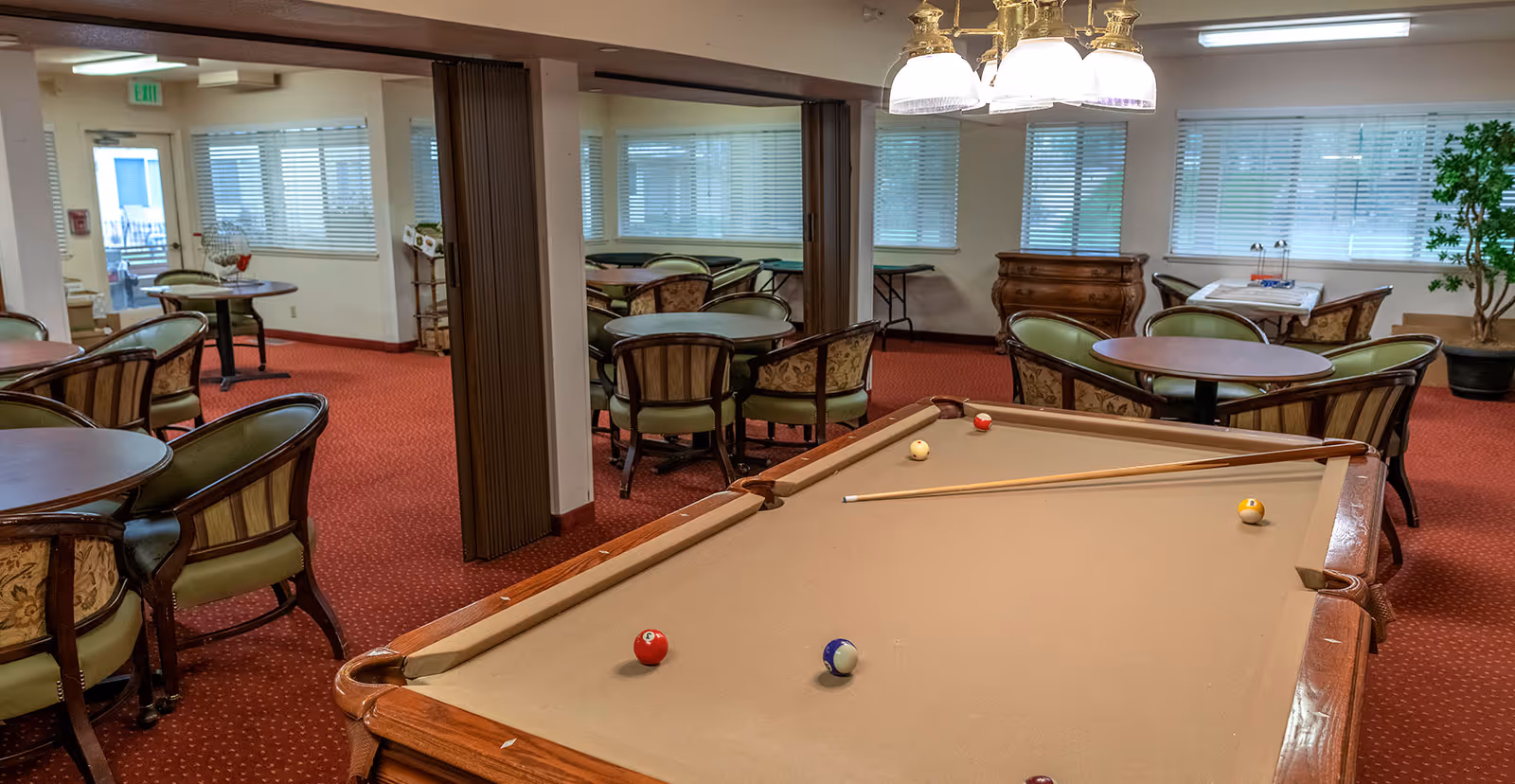 Interior view of a senior living community recreation room featuring a pool table with several billiard balls and a cue stick on it. Surrounding the pool table are multiple round tables with cushioned chairs. The room has red carpet flooring, large windows with blinds, and a chandelier hanging above the pool table. There is a wooden dresser and a potted plant in the background.