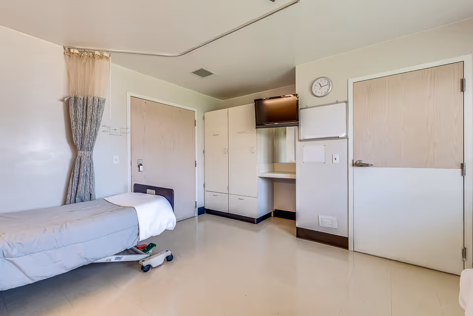 A simple nursing center room with a single hospital bed on wheels, a curtain for privacy, two wooden doors, a wall-mounted TV, a whiteboard, a clock, and built-in cabinets with a small counter area.
