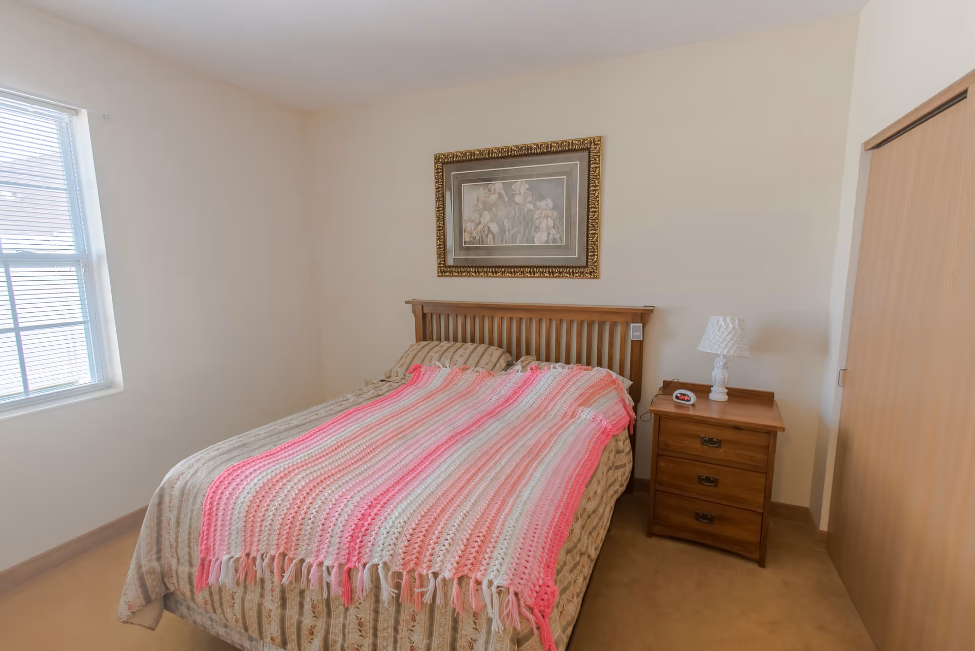 A bedroom with a wooden bed frame and a bed covered with a beige patterned bedspread and a pink and white striped crocheted blanket. There is a wooden nightstand with three drawers next to the bed, holding a white lamp and a digital alarm clock. A framed floral painting hangs on the wall above the bed. A window with blinds is on the left side, and a wooden closet door is on the right.