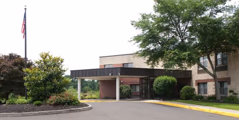 Front entrance of a brick health and rehabilitation center with a covered drop-off, driveway, landscaped beds, and an American flag.