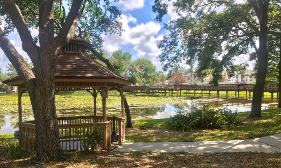 A wooden gazebo surrounded by large trees next to a pond covered with lily pads. A wooden boardwalk extends across the pond, with buildings visible in the background under a partly cloudy sky.