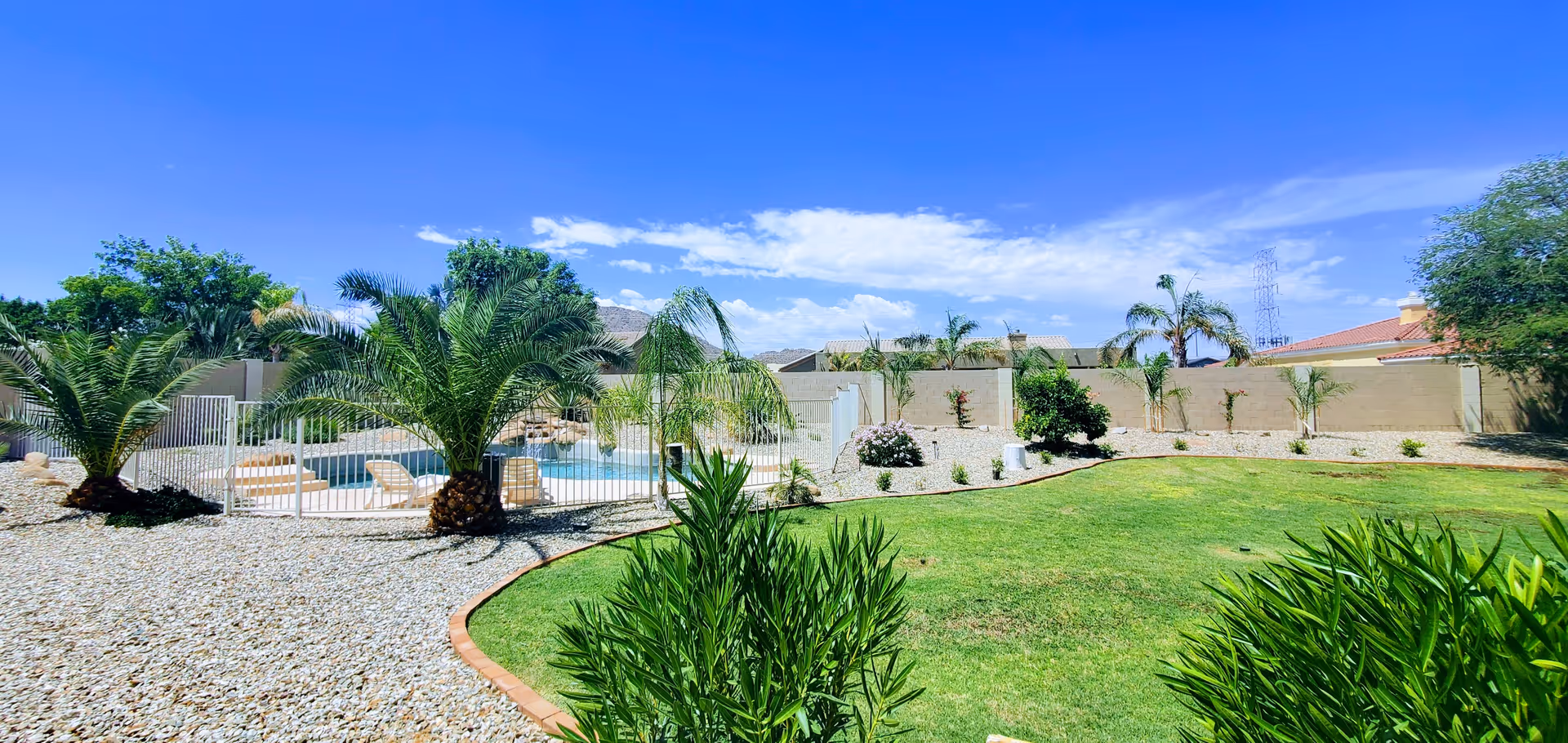 Outdoor area with a fenced swimming pool surrounded by palm trees and lounge chairs, a green lawn, and various shrubs and plants under a bright blue sky with some clouds.