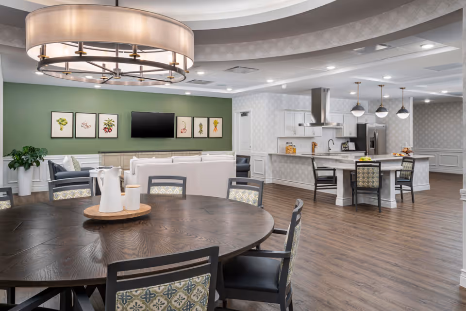 Spacious and modern common area in a senior living facility featuring a large round wooden table with patterned chairs in the foreground, a cozy seating area with white sofas and a wall-mounted TV against a green accent wall with framed botanical prints, and an open kitchen with a large island, pendant lights, and stainless steel appliances in the background.