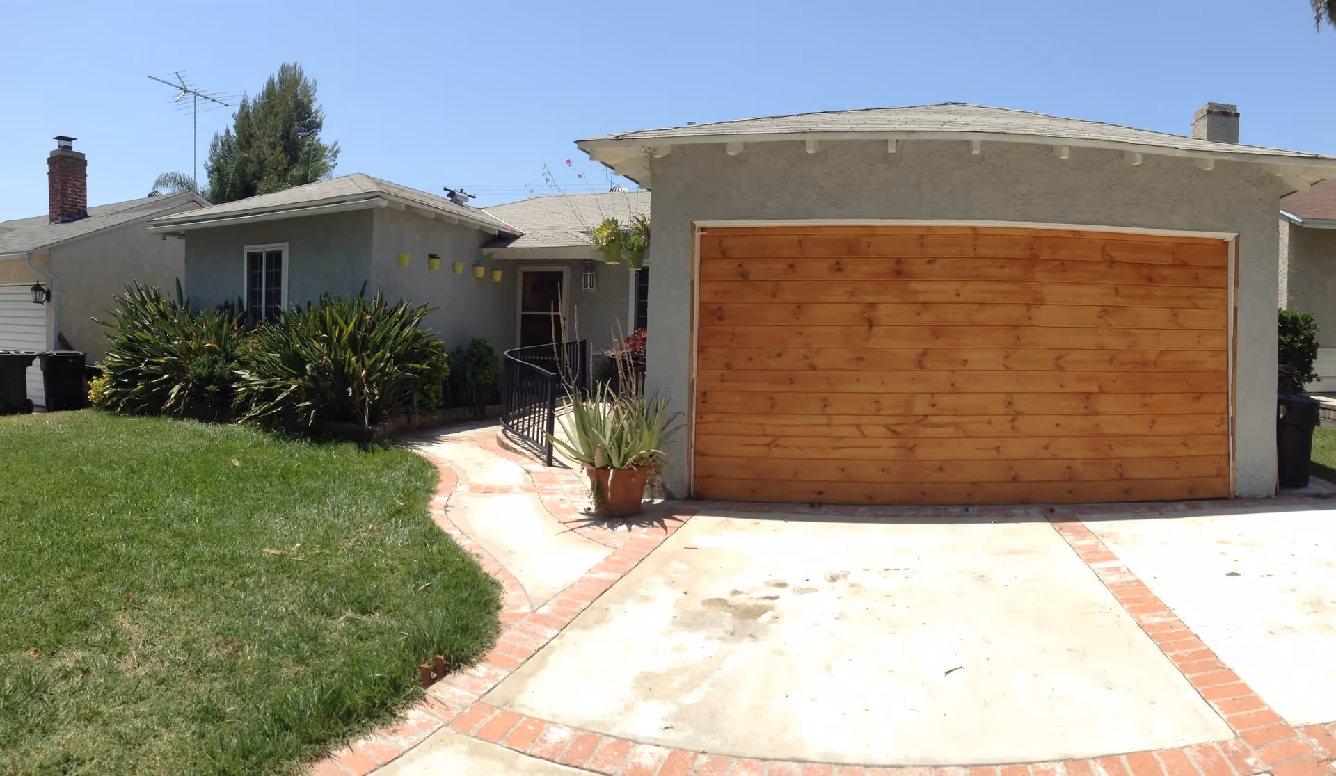 Front exterior view of a single-story house with a wooden garage door, a concrete driveway with brick trim, a curved walkway leading to the front door, green lawn, and various plants and shrubs around the entrance under a clear blue sky.