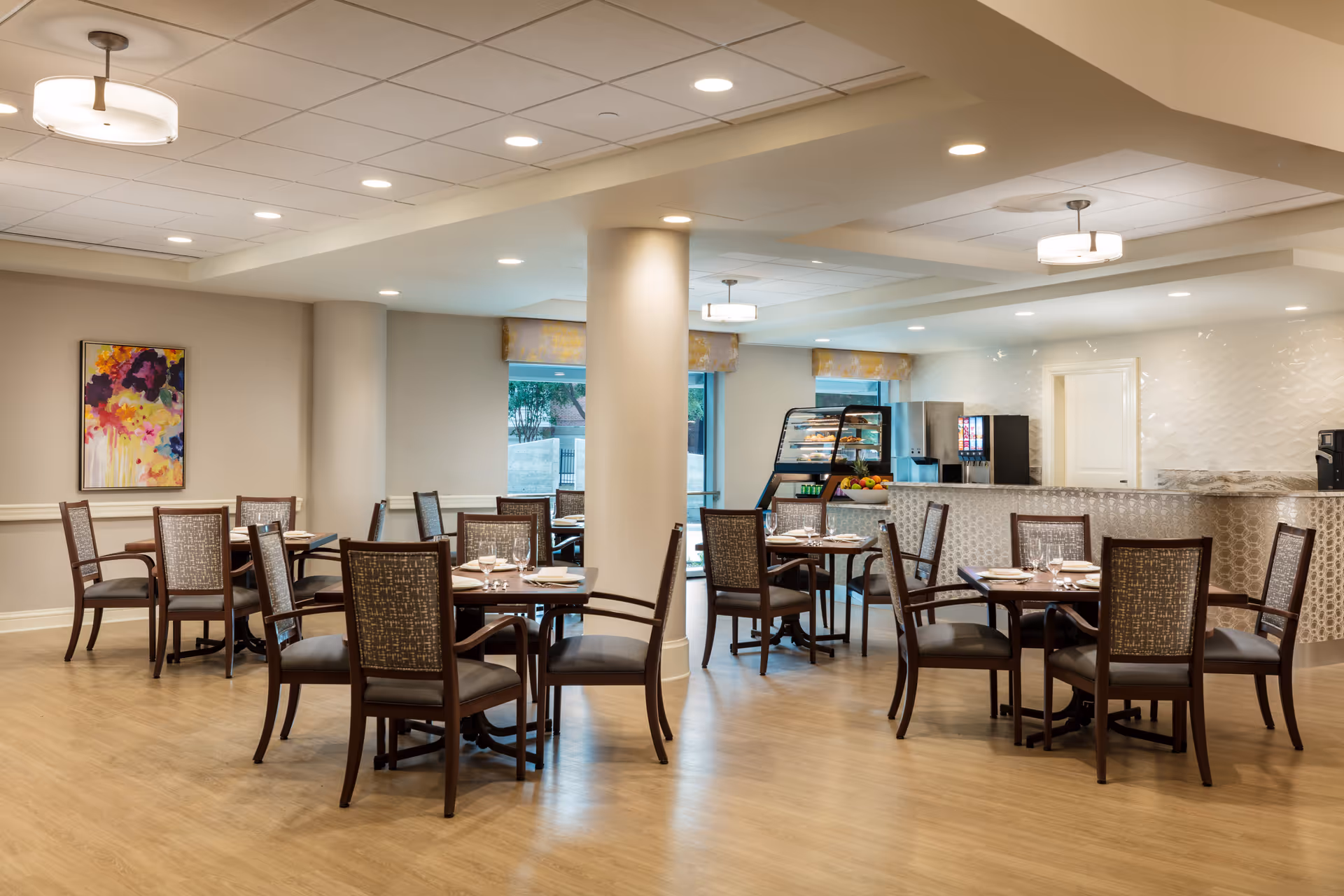 A well-lit dining area with several wooden tables and chairs arranged neatly. Each table is set with plates, napkins, and glassware. The room features light-colored walls, a wooden floor, and ceiling lights. In the background, there is a counter with a display case containing pastries, a soda fountain, and a fruit bowl. Large windows allow natural light to enter the space.