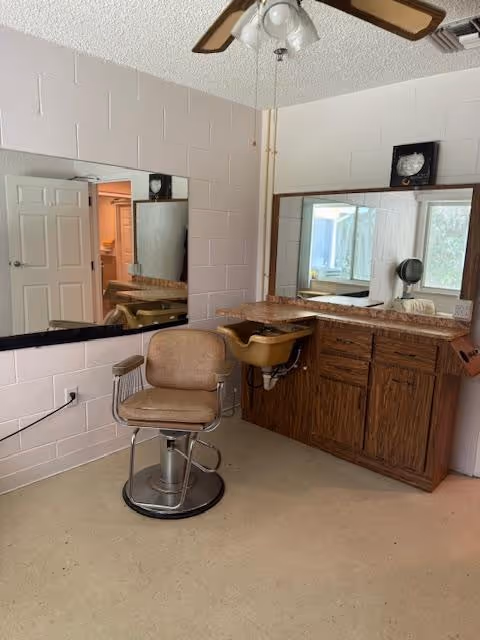 Interior room with a vintage salon chair in front of a large mirror mounted on a wall. There is a wooden cabinet with a countertop and a built-in sink. The walls are painted light pink, and there is a ceiling fan with lights above. A door is open in the background, showing another room.
