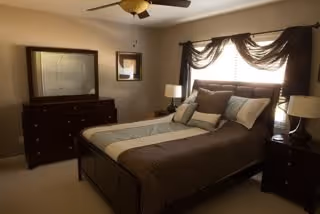 A cozy bedroom with a neatly made bed featuring brown and beige bedding, two bedside tables each with a lamp, a ceiling fan, a dresser with a large mirror, and a small framed picture on the wall. The room is softly lit by natural light coming through a window with dark curtains.