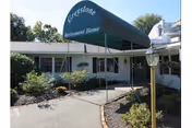 Entrance of Greystone Retirement Home with a green awning displaying the facility name. The building is single-story with white exterior walls, surrounded by shrubs and a paved walkway leading to the door. A lamp post is visible on the right side of the walkway.