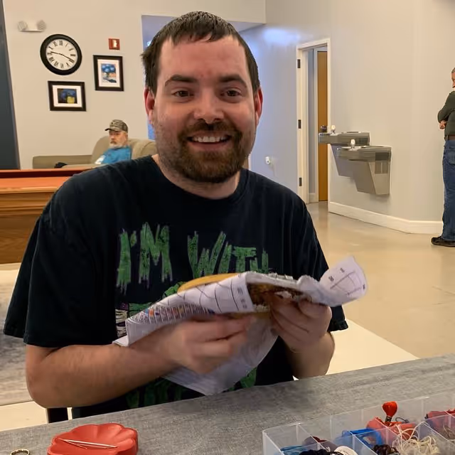 A smiling man with a beard is sitting at a table holding a wrapped sandwich or food item. In the background, there is a man sitting on a couch, a clock on the wall, framed pictures, a water fountain, and another person standing near the wall in a room with light-colored walls and floor.