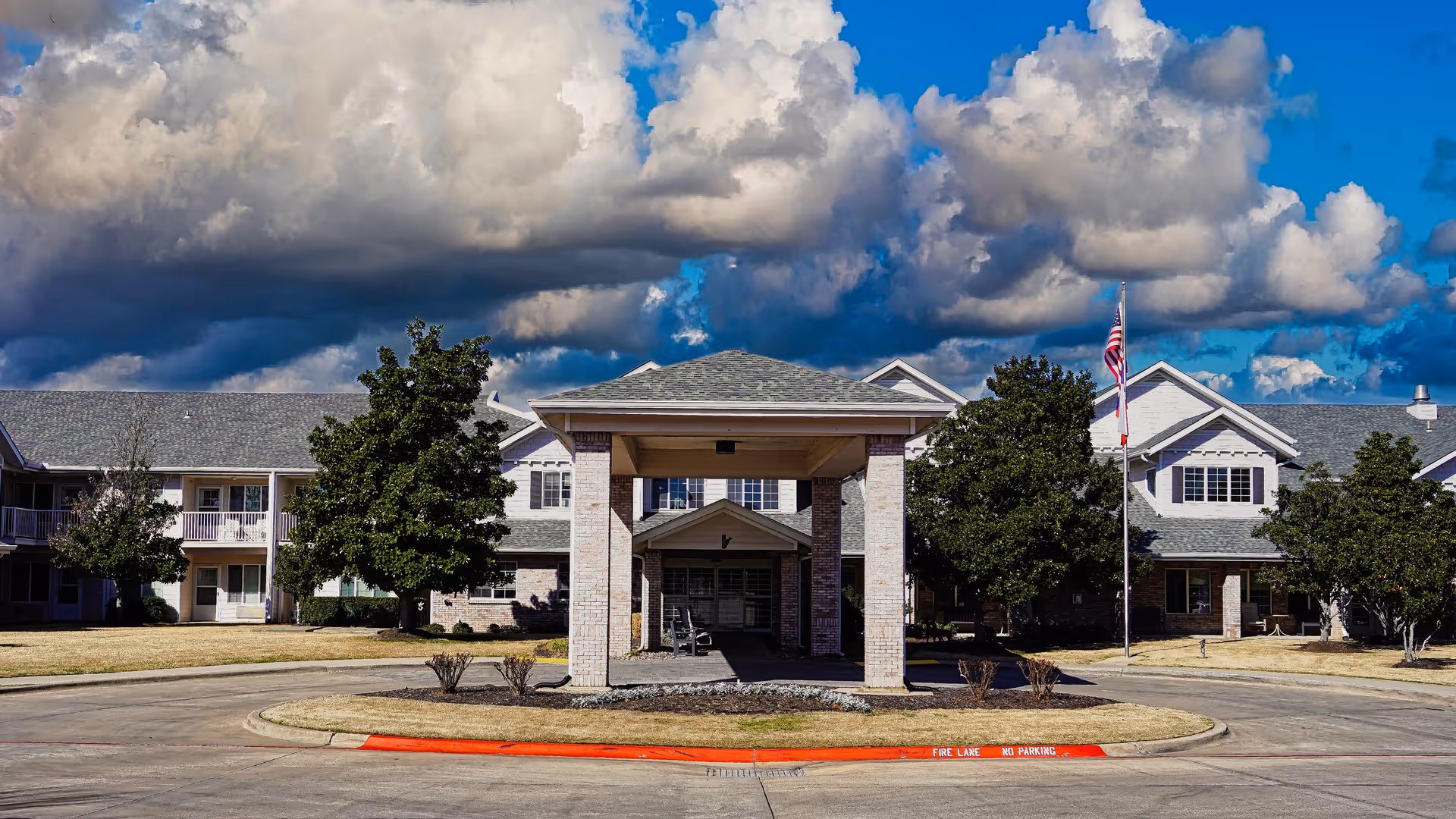 Front exterior view of Pinewood Hills - Sky Active Living facility with a covered entrance, surrounded by trees and a circular driveway under a partly cloudy sky.