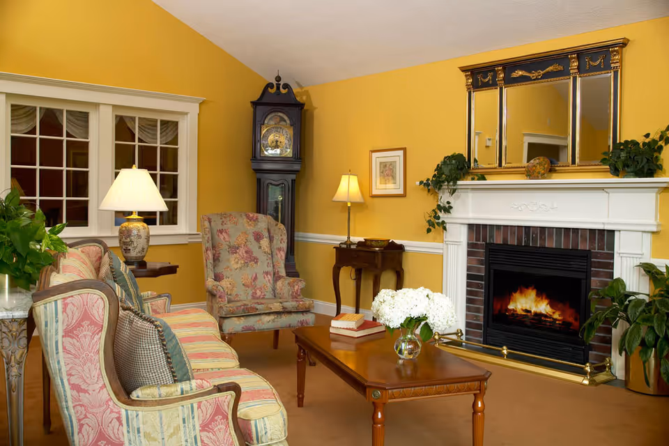 A cozy living room with yellow walls featuring a floral patterned armchair and a striped sofa with cushions. A wooden coffee table with a vase of white flowers and books sits in front of a white fireplace with a burning fire. Above the fireplace is a large ornate mirror with gold accents. There is a tall grandfather clock in the corner and two table lamps providing warm lighting. Several green plants add a touch of nature to the room.