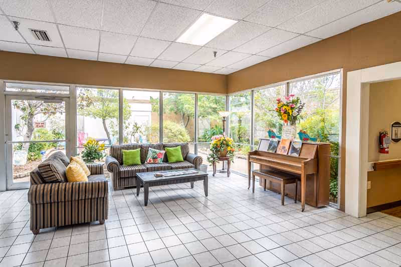 Sunlit common lounge with striped sofas, a coffee table, and an upright piano beside large windows overlooking a garden.